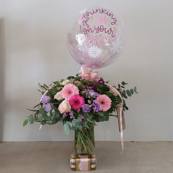 Bouquet of pink, purple, and cream flowers with greenery in a clear vase, topped by a pink-tinted balloon with 'thinking of you' text and a pink ribbon; chocolates at the base.